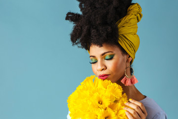 Wall Mural - Portrait of a beautiful young woman  with green eye shadow make up smelling bouquet of fresh yellow daffodil flowers, isolated on blue background