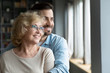© fizkes - Young adult son hugs elderly mother, family smiling looking out the window standing in living room at home. Offspring and future, grown-up child give care to mom, visit her provide attention concept