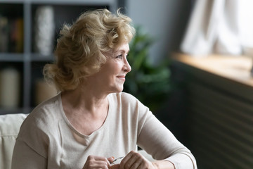 Pretty middle-aged woman sitting on sofa resting at home looking out the window enjoy retired carefree day, old grandma takes off glasses looks at distance daydreams, feels confident in future concept