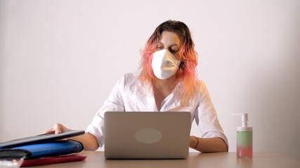 Female nurse in protective medical mask sitting at the table with laptop. Nearby is a liquid soap or sanitizer. Coronavirus or Covid-19.