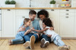 © fizkes - Happy multiracial young parents relax on warm wooden floor in kitchen with little daughters, smiling family with small girls children sit rest in modern design renovated home, have fun together