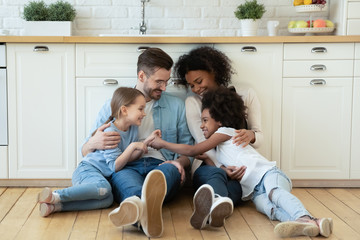 Happy multiracial young parents relax on warm wooden floor in kitchen with little daughters, smiling family with small girls children sit rest in modern design renovated home, have fun together