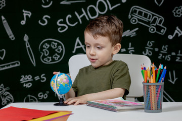 Cute clever boy is sitting at a desk with globe in hand on background with blackboard . Ready for school. Back to school