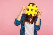 Happy Playful Girl In Checkered Shirt Hiding Behind Yellow Hashtag Symbol, Covering Face With Hash Sign, Concept Of Blogging And Viral Topics On Internet. Studio Shot Isolated On Pink Background