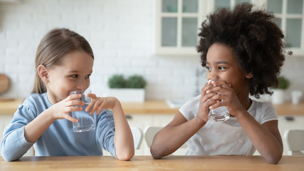 happy little multiracial sisters sit at table in home kitchen drink pure mineral water, smiling smal