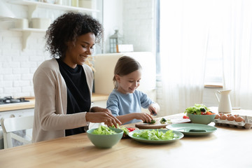 Wall Mural - Happy young biracial mother and little Caucasian daughter chop vegetables prepare healthy salad at home kitchen, smiling African American mom and small girl child cooking making breakfast together