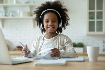 Portrait of smiling little biracial girl in headphones do homework study online in kitchen, happy small African American child in earphones have online web class or lesson using laptop at home