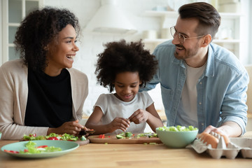 Wall Mural - Happy young multiracial family with little daughter have fun cooking in the kitchen together, smiling multiethnic parents teach small girl child preparing healthy food at home, dieting concept