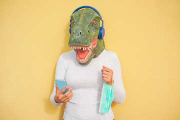 Crazy woman wearing t-rex and holding protection mask while listening music with phone app - Quarantine isolation lifestyle during coronavirus time - Absurd and funny trend concept - Focus on face