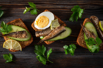 Wall Mural - sandwiches with sprats, fresh cucumber, parsley, lime, rye bread  and boiled egg on a dark wooden background, top view, canned fish,