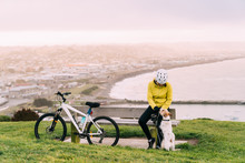 Asian Woman Making Uphill With Mountain Bike. A Woman Playing With A Dog At Lookout Point Oamaru, New Zealand.