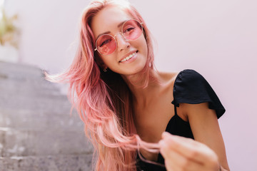 Wall Mural - Easy-going young woman with pink hair posing in summer day. Outdoor photo of smiling dreamy girl sitting on stairs.