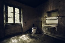 Bathroom With A Sink On The Wall Covered In The Dirt Under The Lights In An Abandoned Building