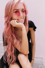 Wall Mural - Pleased good-looking girl in sunglasses touching her face and smiling. Outdoor portrait of cheerful lightly-tanned woman with pink hair posing on white background.