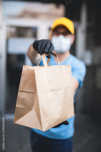 Delivery guy with protective mask and gloves holding box / bag with groceries in front of a building.