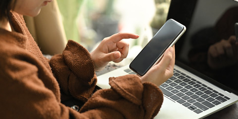 Photo of beautiful woman relaxing on her holidays by using a black blank screen smartphone while sitting in front her black blank screen computer laptop at the leather couch in modern sitting room.