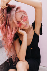 Wall Mural - Glad female model posing with interested face expression on light background. Portrait of magnificent pink-haired woman wears black attire and summer glasses in warm day.