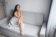 © Алина Троева - Beautiful bored slender brunette woman sitting in comfortable grey couch under warm blanket at home. Self isolation quarantine during Corona virus pandemic. COVID19 stay home save lives concept
