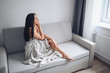 © Алина Троева - Beautiful bored slender brunette woman sitting in comfortable grey couch under warm blanket at home. Self isolation quarantine during Corona virus pandemic. COVID19 stay home save lives concept