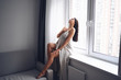 © Алина Троева - Beautiful bored slender brunette woman sitting next to window windowsill under warm grey blanket at home. Self isolation quarantine during Corona virus pandemic. COVID19 stay home save lives concept