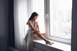 © Алина Троева - Beautiful bored slender brunette woman sitting next to window windowsill under warm grey blanket at home. Self isolation quarantine during Corona virus pandemic. COVID19 stay home save lives concept