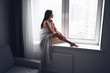 © Алина Троева - Beautiful bored slender brunette woman sitting next to window windowsill under warm grey blanket at home. Self isolation quarantine during Corona virus pandemic. COVID19 stay home save lives concept