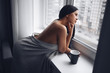 © Алина Троева - Beautiful bored slender brunette woman sitting next to window windowsill under warm grey blanket at home. Self isolation quarantine during Corona virus pandemic. COVID19 stay home save lives concept