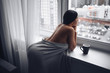 © Алина Троева - Beautiful bored slender brunette woman sitting next to window windowsill under warm grey blanket at home. Self isolation quarantine during Corona virus pandemic. COVID19 stay home save lives concept