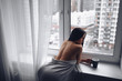 © Алина Троева - Beautiful bored slender brunette woman sitting next to window windowsill under warm grey blanket at home. Self isolation quarantine during Corona virus pandemic. COVID19 stay home save lives concept