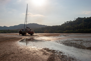 A catamaran with a long mast stands on the sand at low tide against the background of the shore. Sunny. Back light. Horizontal.