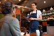 Young friendly waitress with touchpad standing by table with one of clients