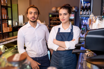 Wall Mural - Two young colleagues in white shirts and denim aprons standing by workplace