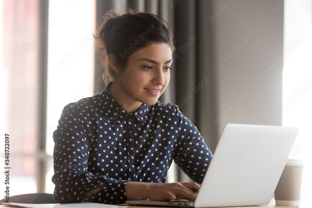 Indian woman sitting at desk in office working on laptop typing business e-mail, check media news, solve current work-related issues distantly, wireless modern tech usage, communication online concept - obrazy, fototapety, plakaty 
