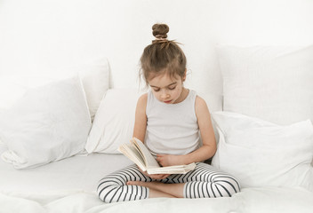 Wall Mural - Cute little girl reading a book on the bed in the bedroom.