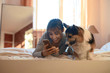 © vitaliymateha - Joyful smiling girl lying on bed in bedroom with her cute Welsh Corgi dog, holding smartphone.