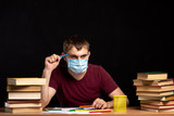 Fototapeta Panele - young man in a medical mask sits at a table with books on a background of a black wall. holding a pencil in his hand near his face. quarantine work. pensive look.