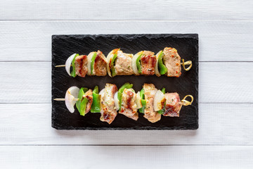 Two portions of grilled pork with onion and green pepper. Barbeque with vegetables, skewers with kebab on black stone plate, on white wooden table, top view.