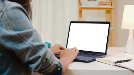 Creative young asia woman sitting at Her desk using laptop with mock up white screen in the cozy living room at modern home.