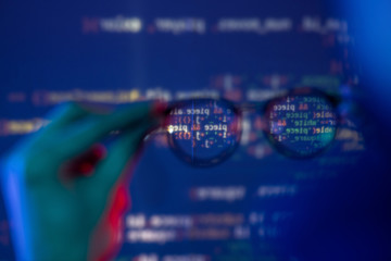 close-up of male hand holding eyeglasses against computer code, defocused background around glasses