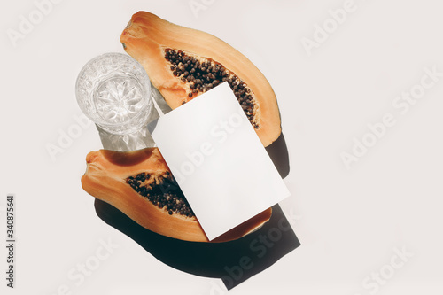 Summer stationery still life scene. Glass of water, cut papaya fruit on white table background in sunlight. Blank paper card, invitation mockup scene. Tropical fruit, vacation concept. Flat lay, top