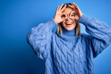 Poster - Young beautiful blonde woman wearing winter wool sweater over blue isolated background doing ok gesture like binoculars sticking tongue out, eyes looking through fingers. Crazy expression.