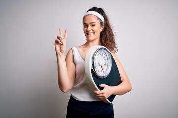 Sticker - Young beautiful woman with curly hair holding weighing machine over white background smiling with happy face winking at the camera doing victory sign with fingers. Number two.