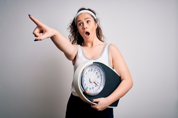 Sticker - Young beautiful woman with curly hair holding weighing machine over white background Pointing with finger surprised ahead, open mouth amazed expression, something on the front