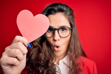 Sticker - Young beautiful woman with curly hair holding paper heart over isolated red background scared in shock with a surprise face, afraid and excited with fear expression