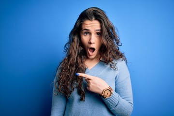 Sticker - Young beautiful woman with curly hair wearing blue casual sweater over isolated background Surprised pointing with finger to the side, open mouth amazed expression.