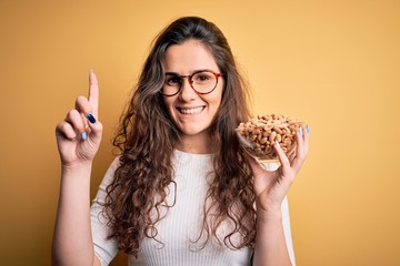 Sticker - Beautiful woman with curly hair holding bowl with healthy peanuts over yellow background surprised with an idea or question pointing finger with happy face, number one