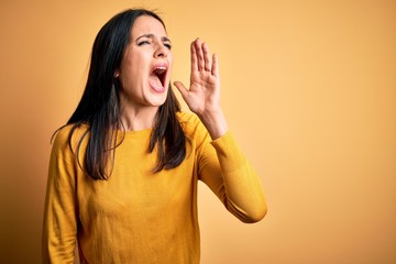 Wall Mural - Young brunette woman with blue eyes wearing casual sweater over yellow background shouting and screaming loud to side with hand on mouth. Communication concept.