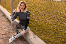Beautiful Woman In Tight-fitting Gray Sportswear Smiling And Looking At The Camera. Sporty Blonde Woman Sitting On The Curb Near The Mesh Fence. Healthy Lifestyle Concept