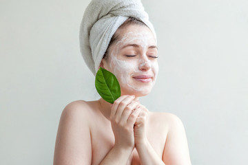 Minimal beauty portrait woman girl in towel on head applying white nourishing mask or creme on face, green leaf in hand isolated white background. Skincare cleansing eco organic cosmetic spa concept.