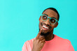 © Carlos David - Portrait of a happy young man with glasses smiling, thinking and looking up with finger on chin, against blue studio background